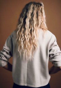 Jasnoszary sweter z teksturowanym wykończeniem, z prążkowanymi mankietami i luźnym krojem, noszony z ciemnymi spodniami, pokazany z tyłu na pomarańczowym tle.