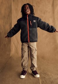 Giacca felpata grigia con accenti neri, zip arancione e logo, abbinata a pantaloni cargo color khaki e sneakers viola. Bambino con le braccia tese.