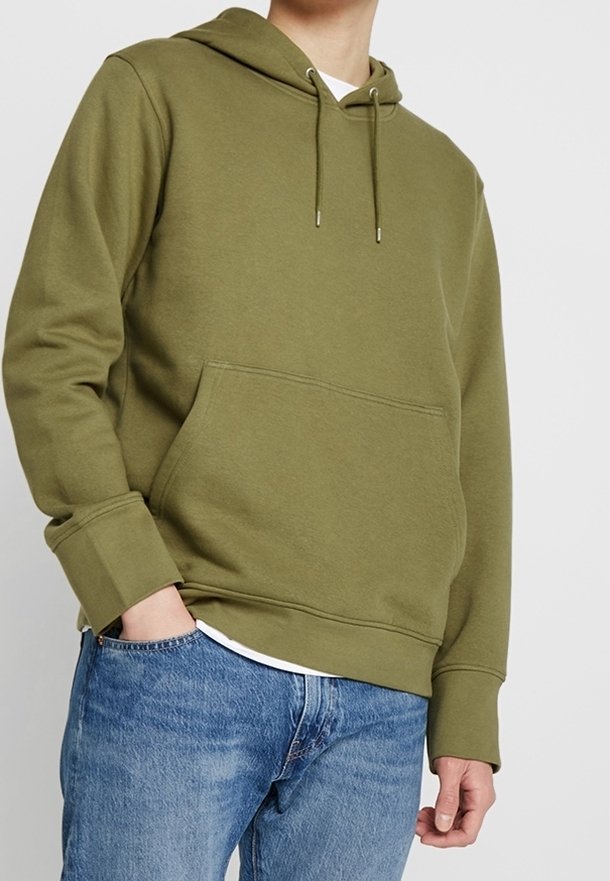 Persona che indossa un felpa con cappuccio verde oliva con tasca a marsupio e jeans blu, in piedi con una mano in tasca.
