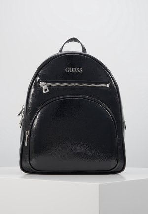 Sac à dos noir brillant Guess avec deux compartiments zippés et une poignée supérieure, présenté sur un piédestal blanc sur fond gris.