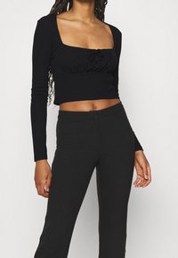 Schwarzes Langarm-Crop-Top mit quadratischem Ausschnitt, gerafftem Vorderteil und tailliertem Design. Kombiniert mit schwarzen, figurbetonten Hosen.
