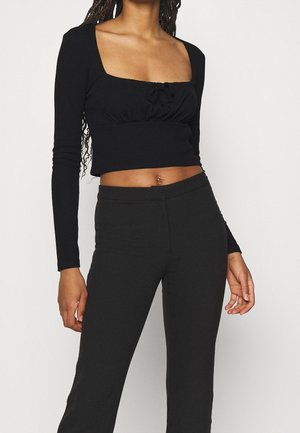 Schwarzes Langarm-Crop-Top mit quadratischem Ausschnitt, gerafftem Vorderteil und tailliertem Design. Kombiniert mit schwarzen, figurbetonten Hosen.