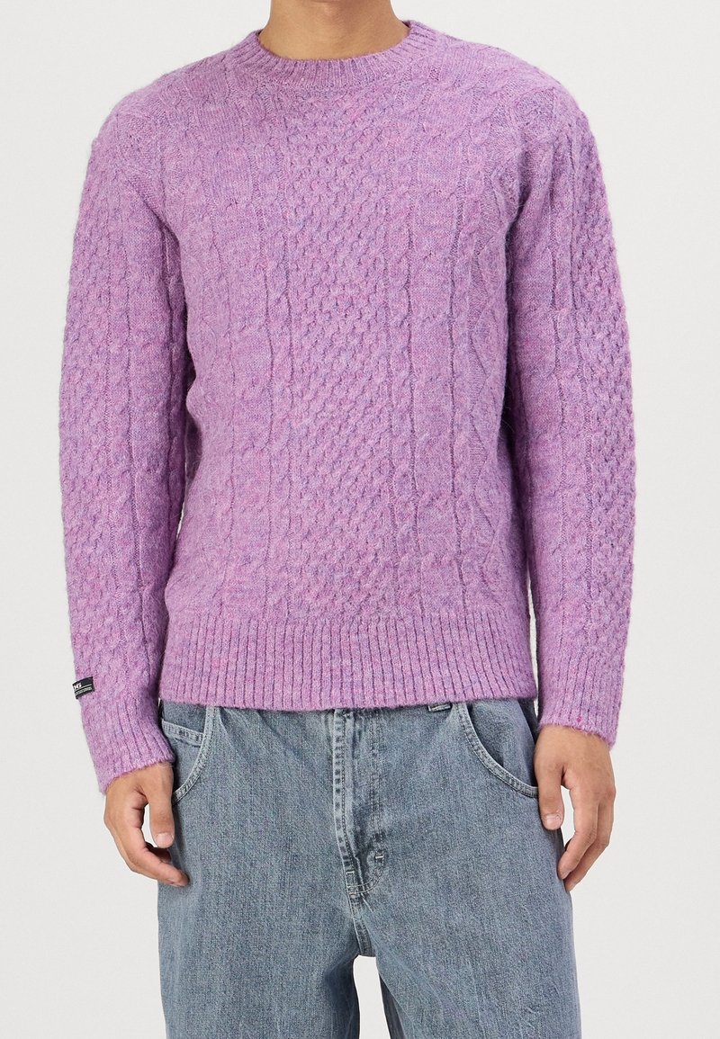 Homme portant un pull en maille torsadée lilas et un jean bleu clair ample, debout devant un fond clair uni.