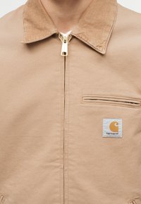 Μπεζ μπουφάν Carhartt με γιακά από κοτλέ, μπροστινό φερμουάρ, τσέπη στο στήθος και επώνυμη ετικέτα στο αριστερό πλευρό.