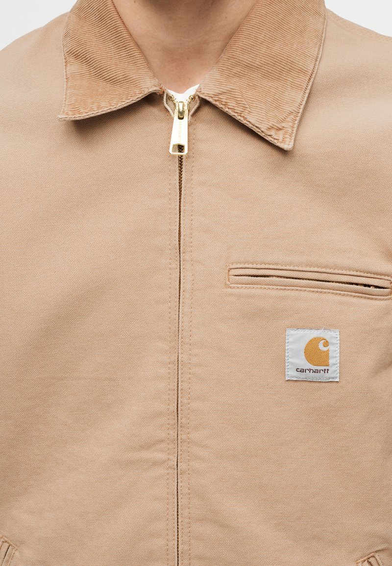 Giacca Carhartt beige con colletto in velluto a coste, cerniera frontale, tasca sul petto e patch con logo marchiato sul lato sinistro.