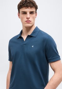 Νεαρός άνδρας που φοράει μπλε πόλο με κοντά μανίκια και μικρό λευκό λογότυπο CK στο στήθος, στέκεται μπροστά σε απλή ανοιχτόχρωμη επιφάνεια.