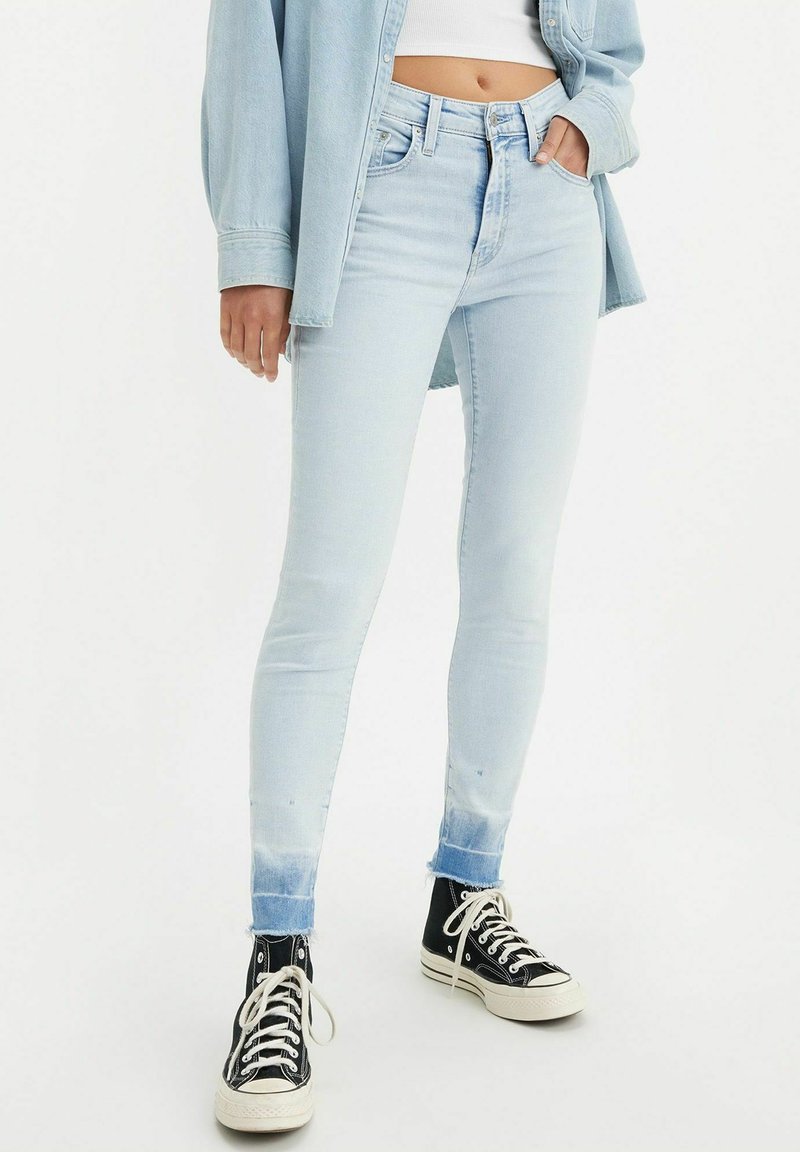 Helle blaue Skinny-Jeans mit einem verblassten Finish und fransigem Saum, kombiniert mit schwarzen High-Top-Sneakern. Das Outfit hat einen lässigen Stil.