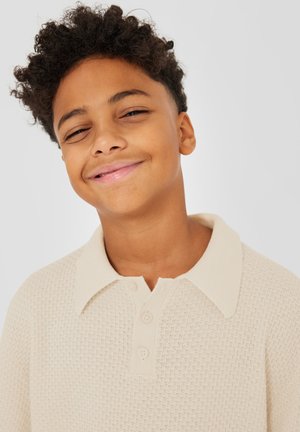 Giovane sorridente con capelli corti e ricci che indossa una polo beige lavorata a maglia con trama, su uno sfondo chiaro e uniforme.