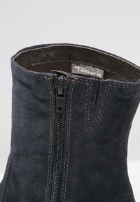 Gros plan d'une bottine cheville en daim noir avec une fermeture éclair noire visible et des coutures marron, montrant l'étiquette de la marque Tamaris à l'intérieur du col.