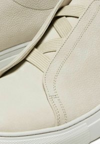 Beige leather sneakers with textured surface, elastic straps, and white rubber soles. Features neat stitching and a minimalist design.