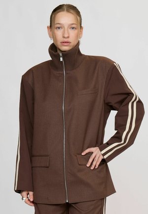 Femme aux cheveux blonds porte une veste marron zippée avec des fines rayures et des bandes beiges sur les manches, posant avec une main sur la hanche.