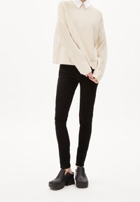 Beige gestrickter Pullover mit oversized Passform, kombiniert mit schwarzen Slim-Fit-Jeans und schwarzen High-Heels mit runder Zehe und chunky Sohle.