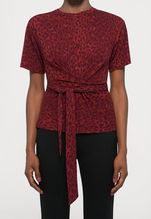 Mujer con una blusa de manga corta con estampado de leopardo en rojo y negro, con cinturón para anudar y pantalones negros sobre un fondo liso.