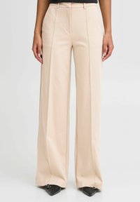 Beige weite Hosen mit glatter Textur, gekennzeichnet durch frontale Falten, eine hohe Taille und einen maßgeschneiderten Schnitt. Kombiniert mit spitzen schwarzen Schuhen.