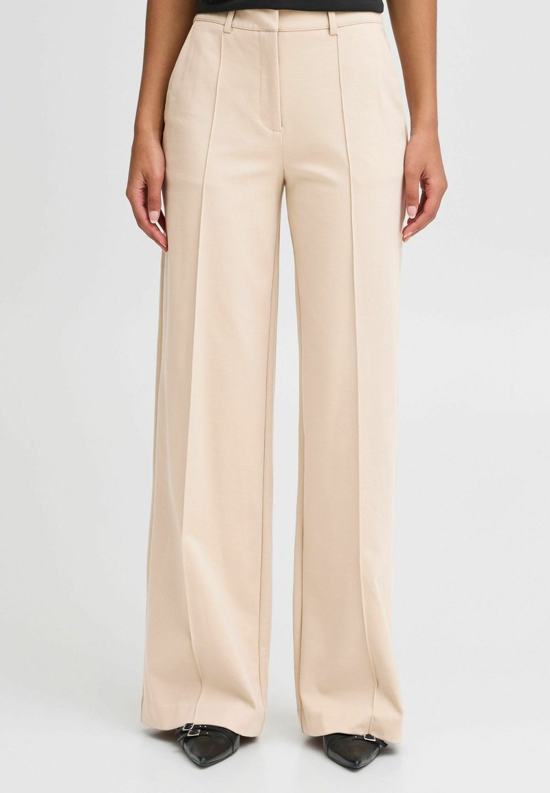 Beige wide-leg trousers with a smooth texture, featuring front pleats, a high waist, and a tailored fit. Paired with pointed black shoes.