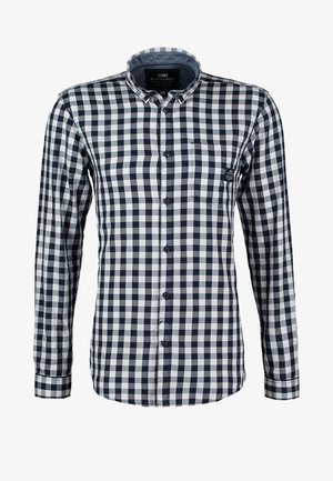 Uomo che cammina indossando una camicia a quadri bianca e nera, jeans grigi, scarpe da ginnastica e porta una borsa a tracolla nera con tracolla marrone.