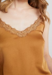 Camiseta de satén mostaza con escote en V adornado con un intrincado acabado de encaje. Textura suave con tirantes ajustables y un corte relajado.