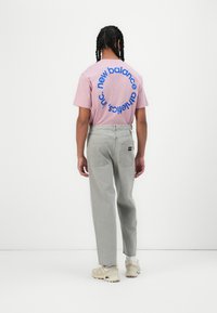 Pink short-sleeve t-shirt with blue circular branding on the back, paired with light gray pants. Feet in neutral-colored sneakers.