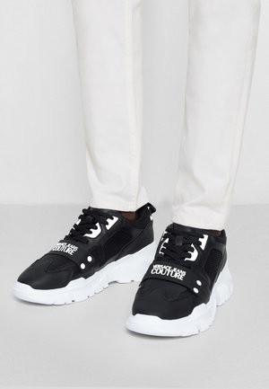 Zapatillas Versace Jeans Couture en blanco y negro, usadas con pantalones blancos, con suelas gruesas y tiras con marca en la parte superior.