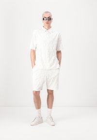 White textured polo shirt and shorts made of soft fabric, featuring an embossed pattern. Model wears oversized silver sunglasses and white sneakers.
