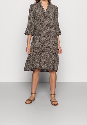 Femme portant une robe noire et blanche à motif longueur genou avec manches trois-quarts et des sandales marron à bride à la cheville, debout sur un sol gris.