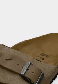 Καφέ σουέντ σανδάλι με φαρδιά λουρί, μεταλλική αγκράφα και εσωτερική επένδυση με καμπύλες. Διαθέτει υφή στην επιφάνεια και ανάγλυφες λεπτομέρειες του λογότυπου.