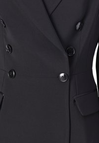 Blazer preto de dupla abotoadura feito de um tecido liso, apresentando botões pretos brilhantes e bolsos laterais com um design limpo e estruturado.