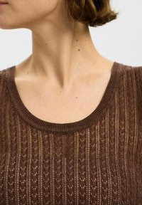 Smeđa pletena majica s izrezom u obliku kruške, koja ima teksturirani uzorak vertikalnih pruga i detalje s otvorenim tkanjem. Mekani materijal.