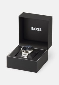 Sudraba nerūsējošā tērauda pulkstenis ar melnu ciparnīcu, ietverts melnā kastē, kurā balti uzrakstīts zīmola nosaukums "BOSS". Maiga spilvena iekšā.