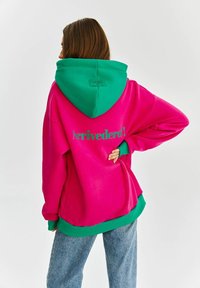 Różowa polarowa bluza z kapturem w zielonym kolorze, z napisem "Arrivederci!" w zielonym na plecach. Luźny krój z ściągaczami przy mankietach i u dołu.