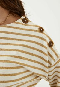 Weißer Pullover mit goldenen horizontalen Streifen, ausgestattet mit drei dekorativen braunen Knöpfen an der Schulter und einem weichen, strukturierten Stoff.