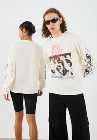 Beige long-sleeve shirt with black graphic print and text on front; black abstract design on sleeves; styled with black shorts and sunglasses.
