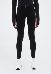 Μαύρο κολάν από ελαστικό υλικό, με εφαρμοστό σχέδιο, λεία υφή και ελάχιστες λεπτομέρειες. Συνδυάζεται με λευκά αθλητικά παπούτσια.