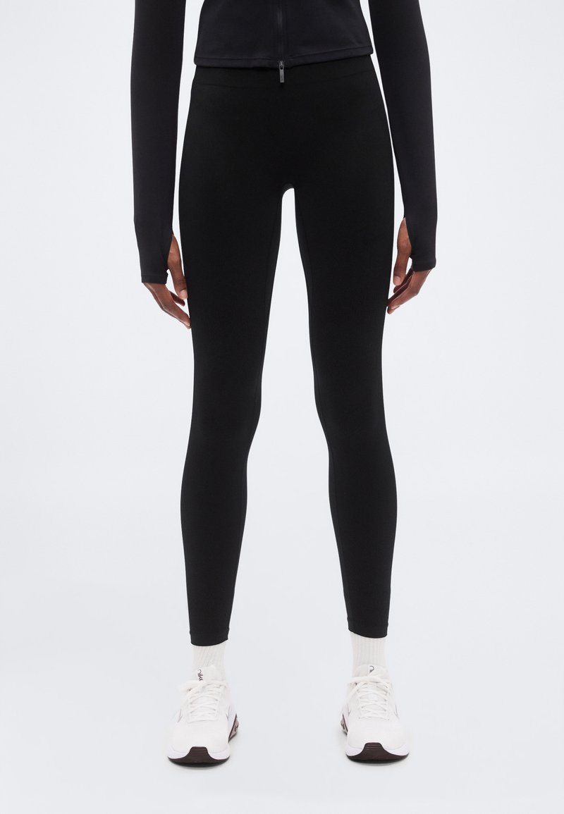 Μαύρο κολάν από ελαστικό υλικό, με εφαρμοστό σχέδιο, λεία υφή και ελάχιστες λεπτομέρειες. Συνδυάζεται με λευκά αθλητικά παπούτσια.