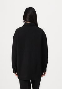 Zwarte oversized shirt gemaakt van stevige stof. Heeft een puntkraag, knoopmanchetten en een relaxte pasvorm met verlaagde schouders.