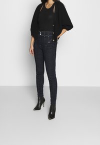 Cardigan de punto negro sobre una camiseta de tirantes negra, combinado con jeans skinny oscuros y botines de tacón alto negros. Botones y detalles de costura visibles.