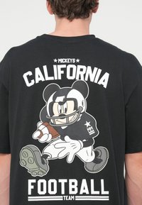 Μαύρο t-shirt με καρτούν χαρακτήρα σε στολή ποδοσφαίρου, κρατώντας μια μπάλα ποδοσφαίρου. Το κείμενο κάτω γράφει «Η ομάδα ποδοσφαίρου της Καλιφόρνια του Μίκι».