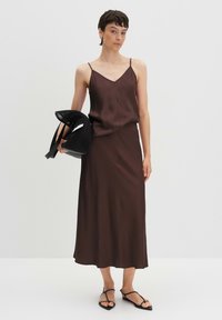 Bruine satijnen camisole en bijpassende midi-rok met dunne bandjes. Zwarte geometrische tas met twee handvatten. Model draagt zwarte sandaaltjes met bandjes.