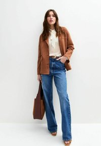 Brown suede jacket, cream blouse with a neck tie, high-waisted blue jeans, brown belt, and a brown tote bag. Sandals on feet.