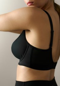 Zwarte sportbeha met een gemêleerd gaaspaneel, verstelbare schouderbanden en een soepele stof. Het ontwerp legt de nadruk op comfort en ondersteuning.