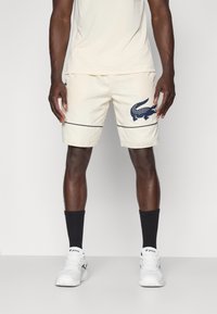 Beige shorts with a navy crocodile logo, contrasting black stripe, and a lightweight material. Worn with black socks and white sneakers.