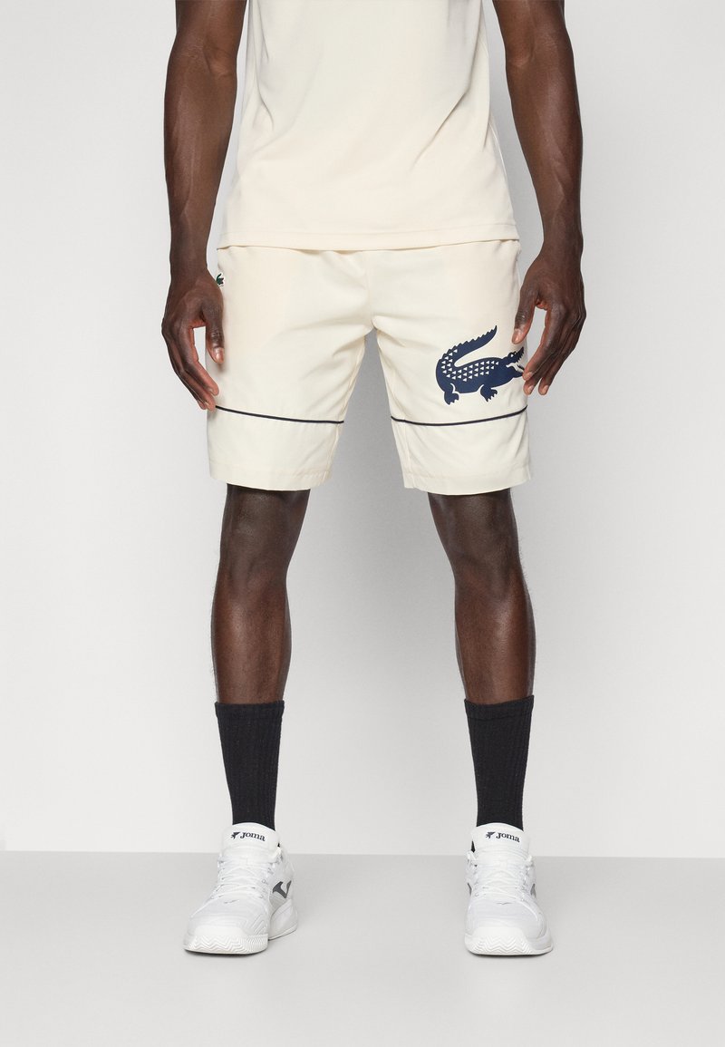 Shorts beiges avec un logo crocodile marine, une bande noire contrastante et un matériau léger. Portés avec des chaussettes noires et des baskets blanches.