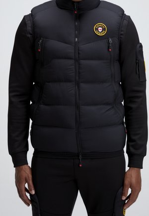 Homme portant une doudoune noire sans manches par-dessus une chemise noire à manches longues et un pantalon noir, avec un patch circulaire de marque sur la poitrine de la doudoune.