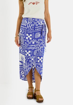 Femme portant un haut blanc sans manches avec une broderie de créatures marines et une jupe portefeuille bleue à motifs, associée à des sandales compensées marron.