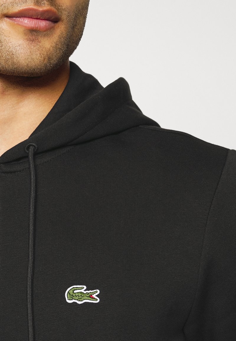 Gros plan d'une personne portant un sweat à capuche noir avec un logo crocodile vert brodé sur la poitrine.