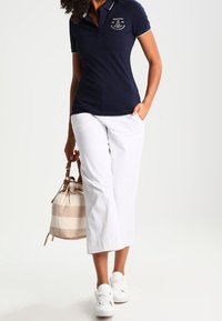 Polo shirt navy avec des rayures blanches sur le col, associé à un pantalon large blanc et des baskets blanches. Tenant un sac rayé beige et crème.