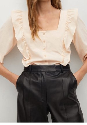 Femme portant un chemisier crème à encolure carrée froncée avec des boutons, associé à un pantalon en cuir noir taille haute, les mains dans les poches.