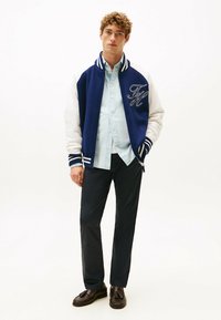 Marineblå og hvid varsity-jakke med broderede initialer, ribbede manchetter og krave, parret med en lyseblå skjorte og mørke bukser.