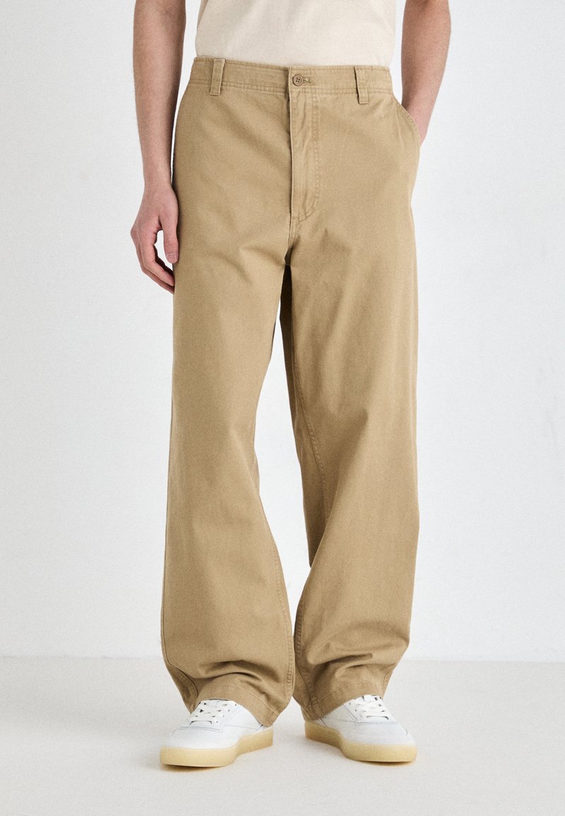 Khakihosen mit einem geraden Schnitt, aus robustem Baumwollstoff, mit Seitentaschen und Knopfverschluss. Getragen mit weißen Sneakers.