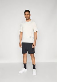 Világos bézs rövid ujjú ing logóval, fekete sportos rövidnadrággal párosítva. Fehér tornacipő és fekete zokni egészíti ki az öltözéket.